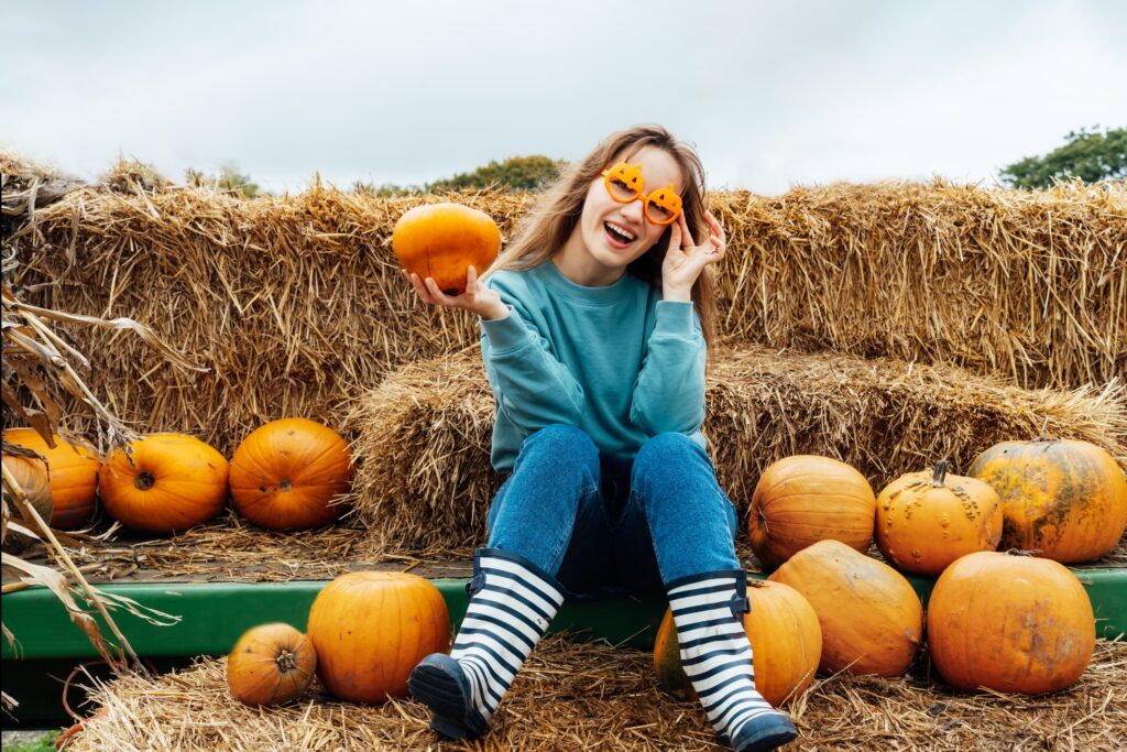 Young woman wearing pumpkin-shaped sunglasses, sitting on hay bales surrounded by pumpkins, celebrating fall festivities in Fayetteville, highlighting community events and family fun, relevant to Storms Orthodontics' local engagement.